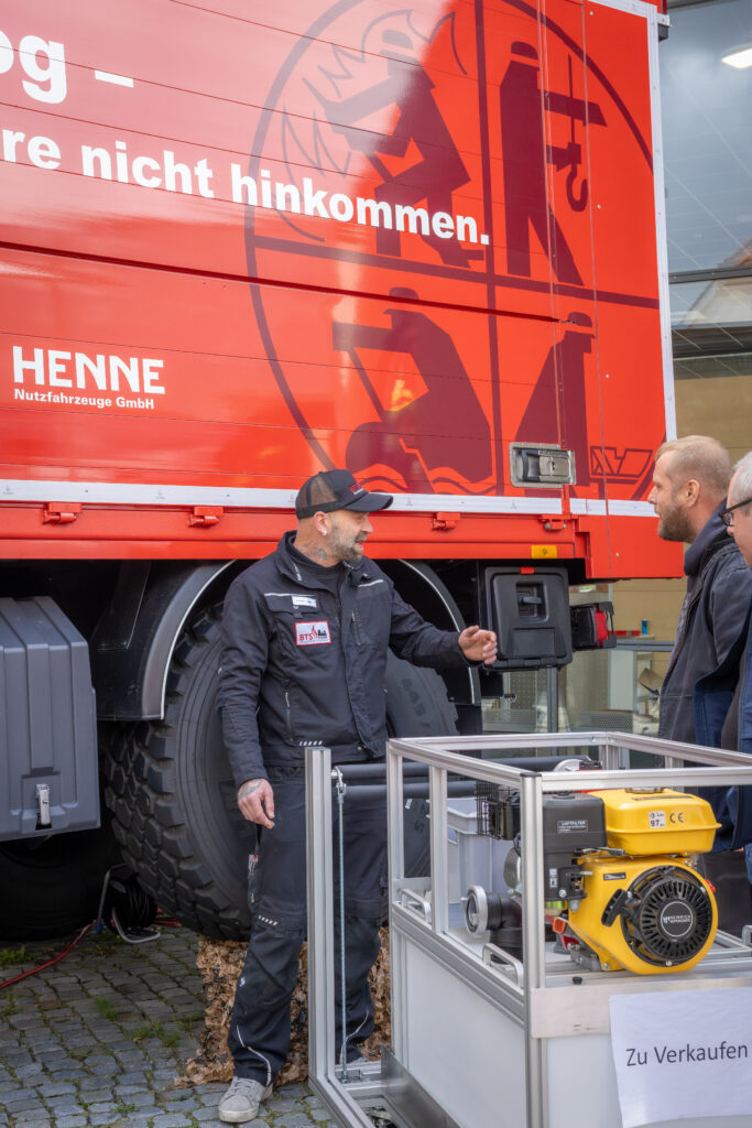 Mitarbeiter erklärt Besuchern eine tragbare Pumpe vor einem roten Unimog-Lkw