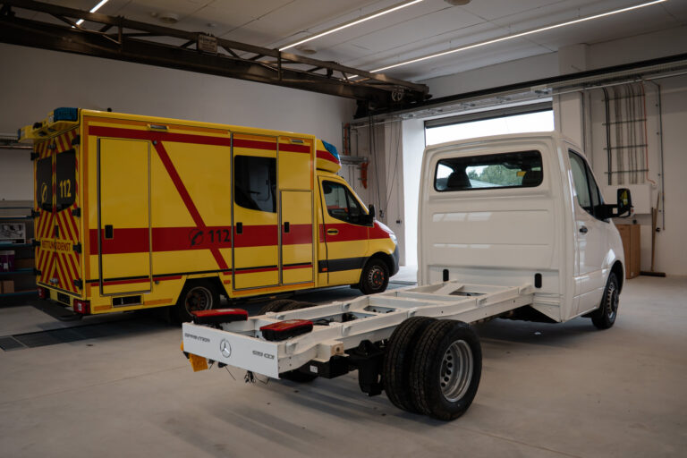 Fahrgestell eines Mercedes Sprinter neben einem gelben Rettungswagen in der Halle