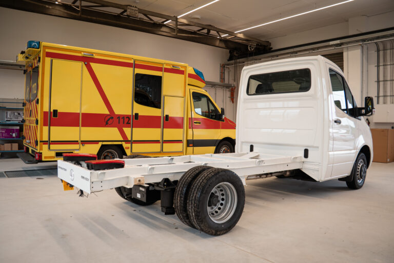 Fahrgestell eines Mercedes Sprinter neben einem fertig ausgebauten gelben Rettungswagen